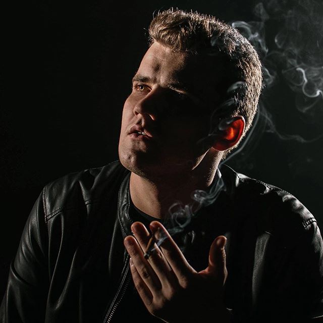 Portrait of a man in a leather jacket smoking a cigarette in a moody, shadowed studio setting.