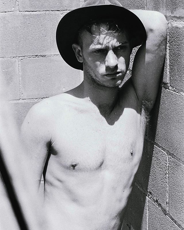 A shirtless man in a hat leans against a brick wall in this black and white portrait.