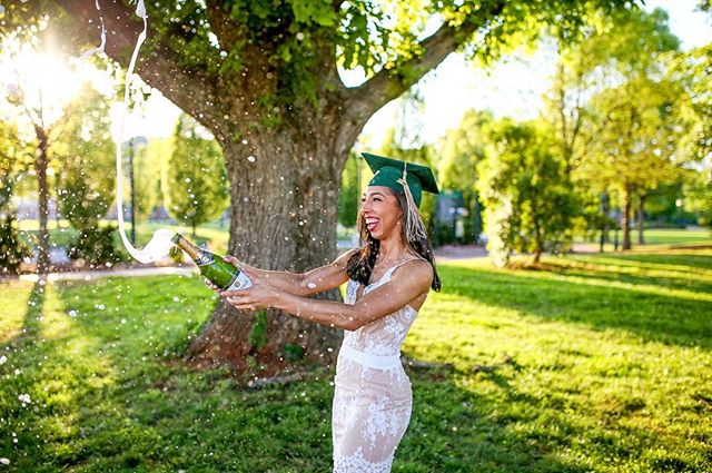 A smiling graduate in a cap pops champagne in a park, celebrating graduation day with joy and excitement.