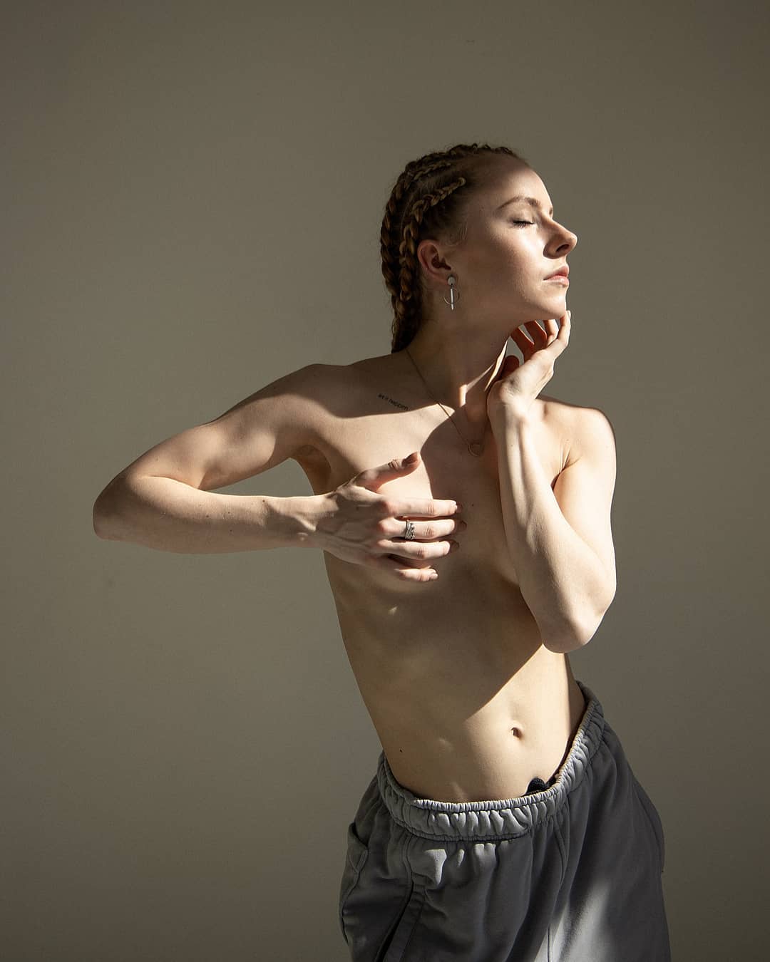 A topless woman in sweatpants poses in a studio with soft, natural light highlighting her form.
