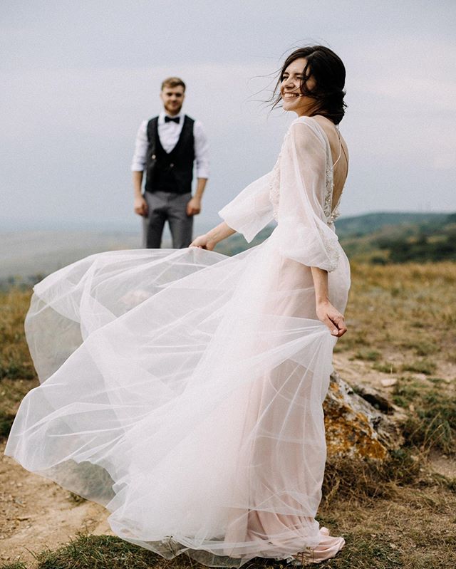 A bride in a flowing wedding dress smiles as her groom stands in the background, captured in a romantic outdoor setting.