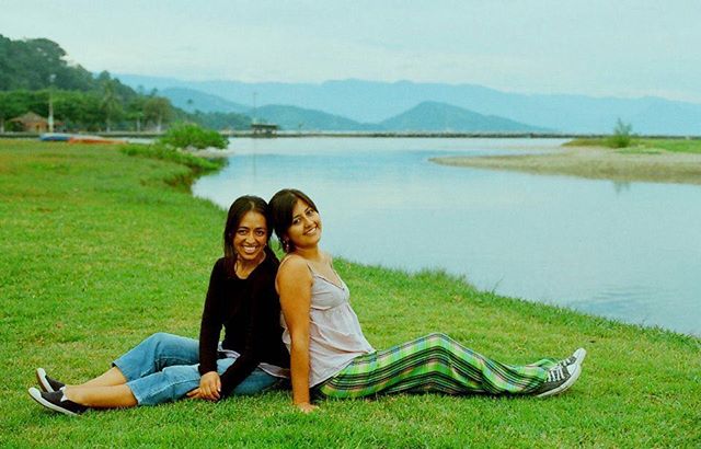 Two happy women sit back-to-back on green grass near a lake, smiling on a cloudy day.