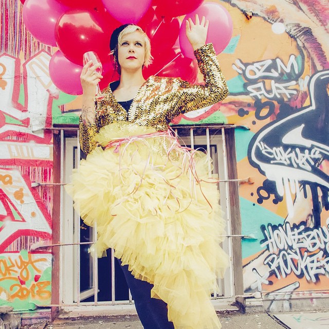 A woman in a yellow tutu and gold jacket celebrates with red balloons and a champagne glass against a graffiti wall.
