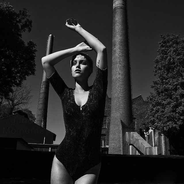 A strong woman confidently poses against an industrial backdrop in a striking monochrome fashion shot.