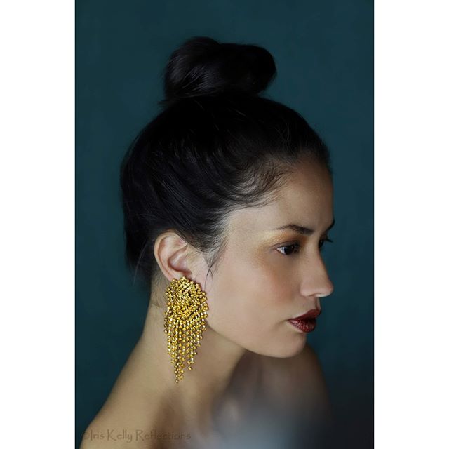 A woman with gold earrings and a bun hairstyle poses against a dark teal background.