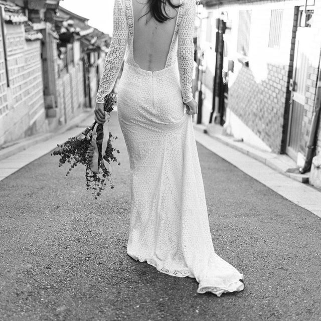 A bride in a lace wedding dress walks down a quiet street, holding a bouquet.