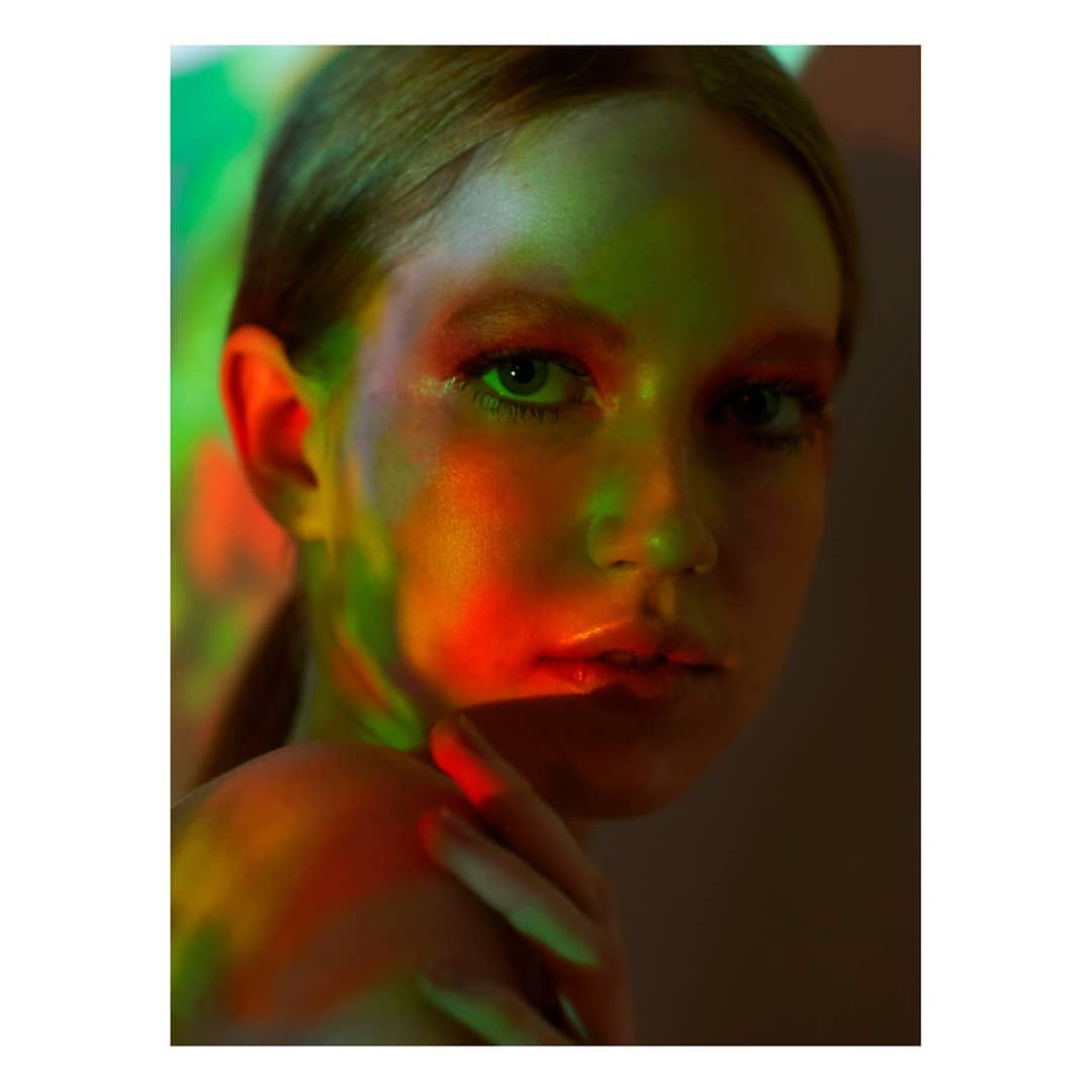 A woman with creative makeup is illuminated by red and green lighting in a studio setting.