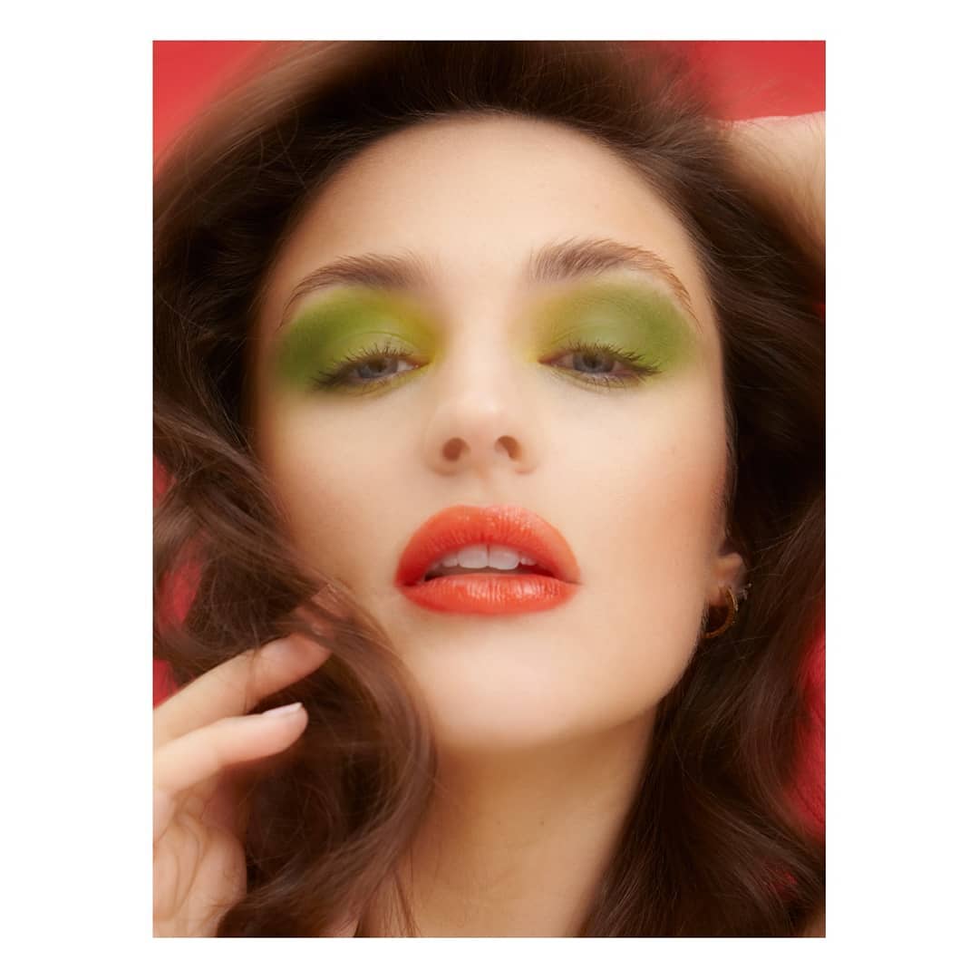 A woman with green eyeshadow and orange lipstick poses for a beauty shot.