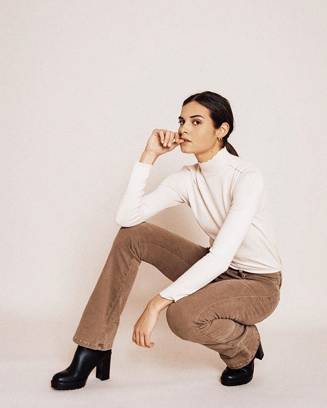 A young woman poses in earth-tone clothing for a minimalist fashion shoot in a studio setting.