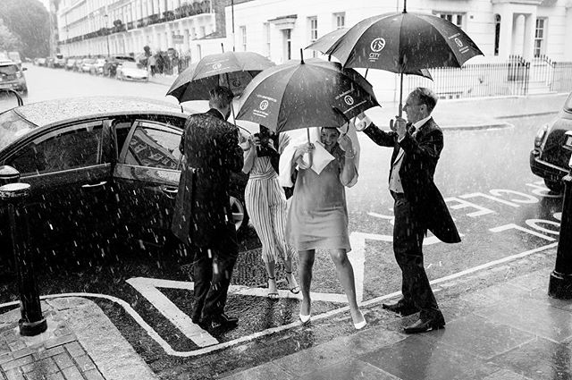 People with umbrellas are assisted from a car during a rainy day in a city.