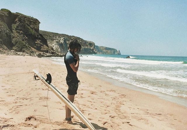 A surfer stands on the beach with his surfboard looking out at the ocean on a sunny day.
