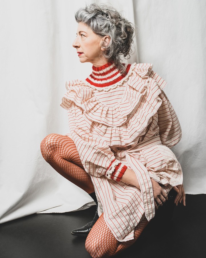 A mature woman with gray hair poses in a vintage-style striped dress and fishnet stockings.