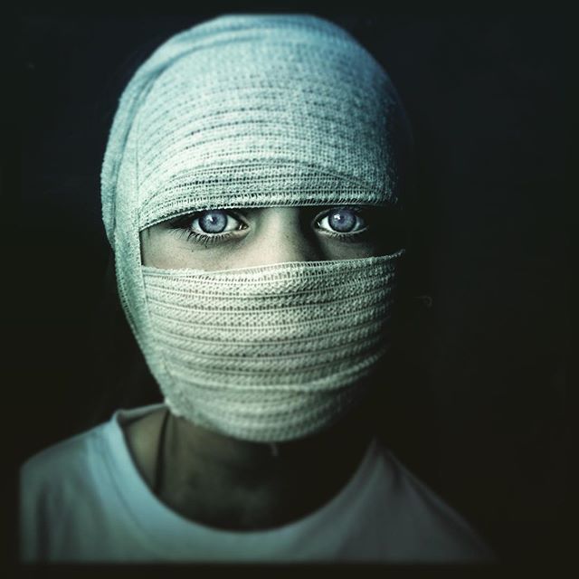 A person with piercing eyes wrapped in bandages stares intensely in a dramatic, close-up studio portrait.
