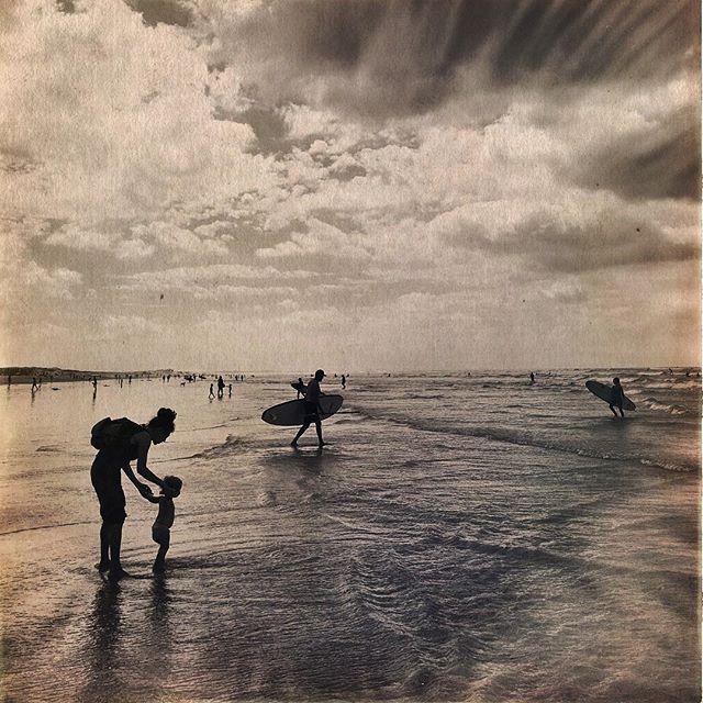 A vintage-style photo captures a day at the beach with people enjoying the ocean and surfers heading out to catch some waves.