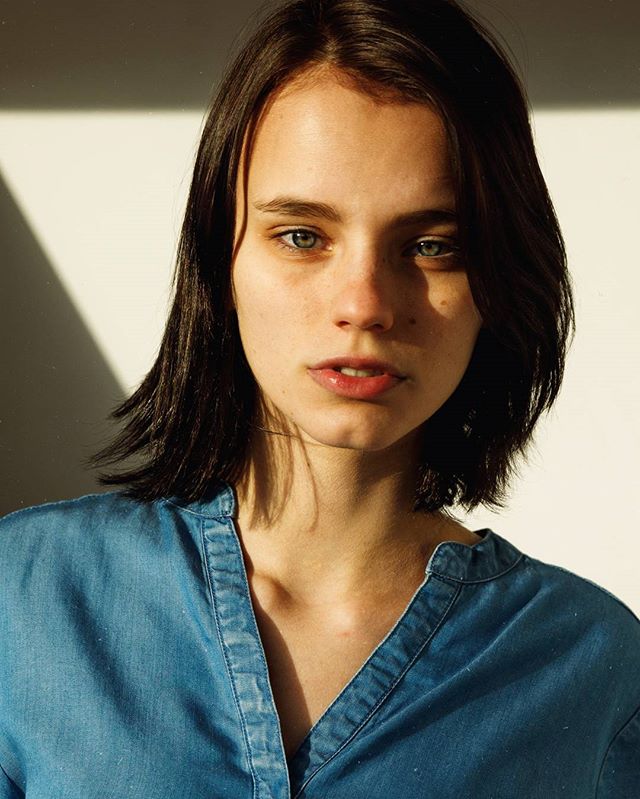 A serene young woman with short brown hair poses in a bright, soft light filled studio for a beauty campaign.