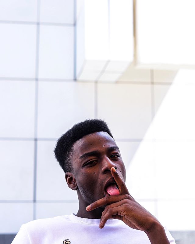 A young man playfully poses with his tongue out in front of a textured white wall on a sunny day.
