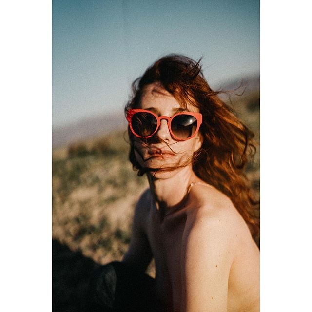 A redhead woman wears red sunglasses in a sun-drenched outdoor scene, portraying calm and relaxed summer vibes.
