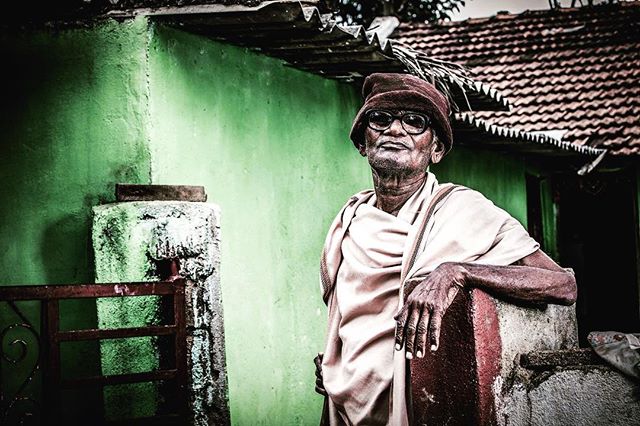 Portrait of an elderly man in a hat and glasses standing in front of a green building.