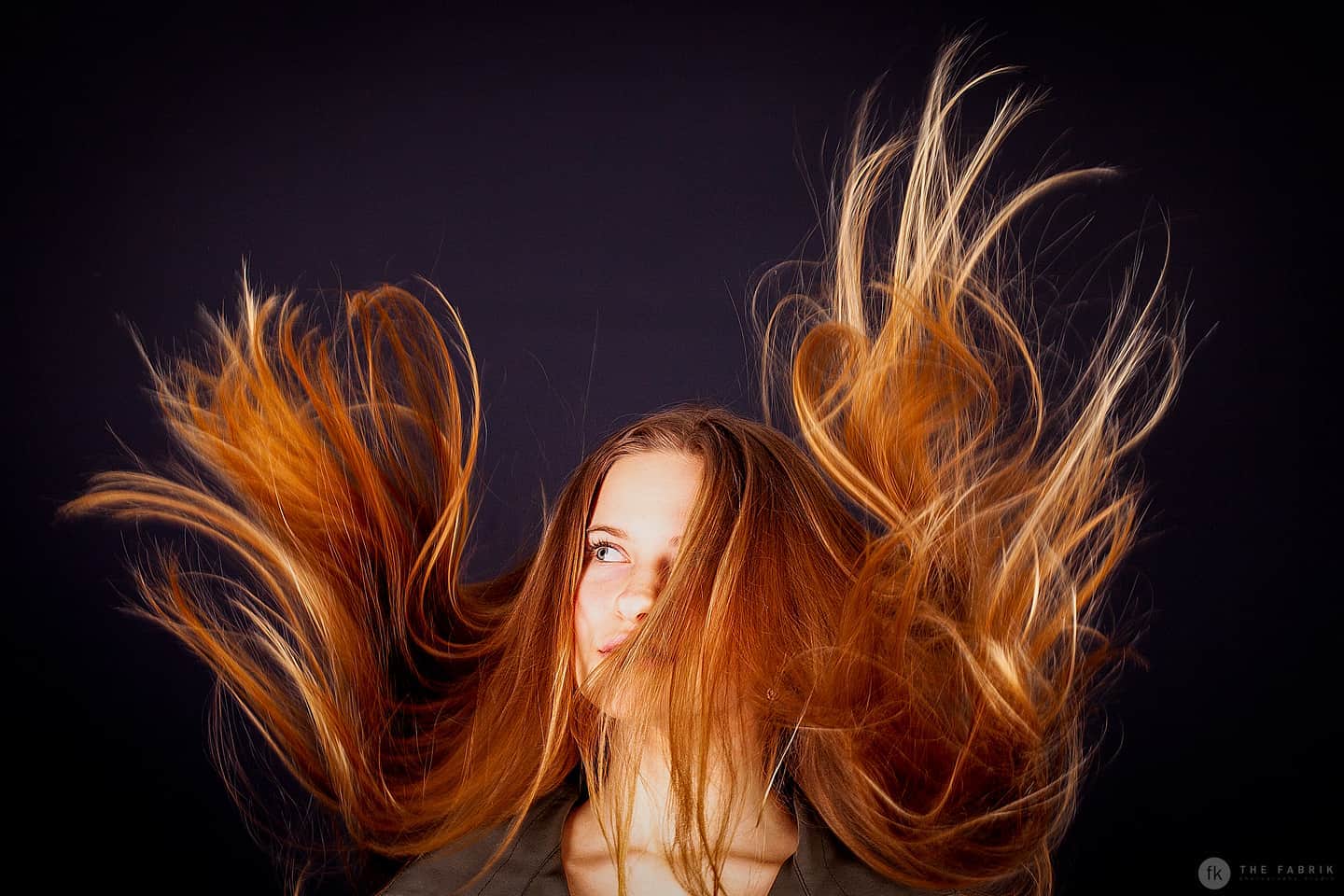 A woman tosses her auburn hair in a dynamic pose against a dark background, creating a sense of energy and movement.