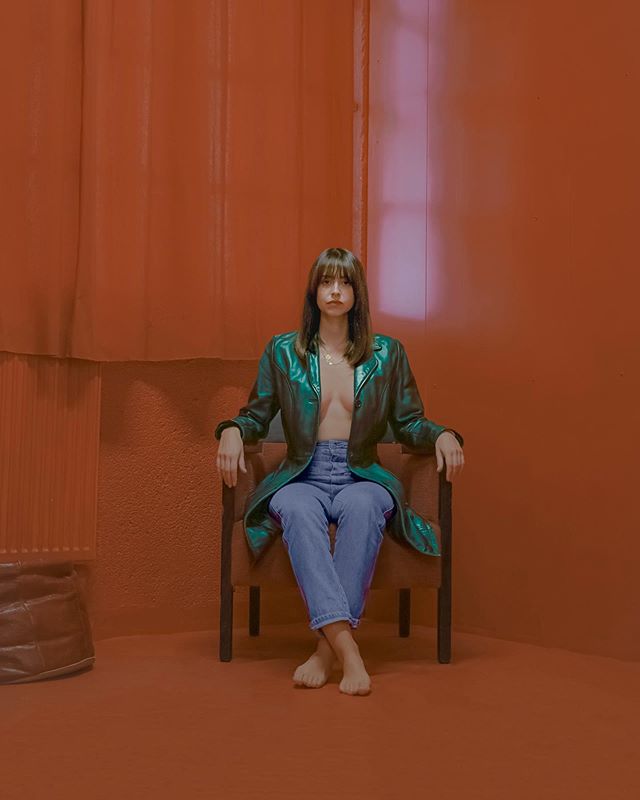 A brunette woman sits in a chair wearing a jacket and jeans in a warm-toned room.