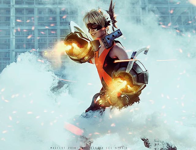 A cosplayer as Bakugo Katsuki strikes a heroic pose with fiery effects in a dynamic outdoor setting.