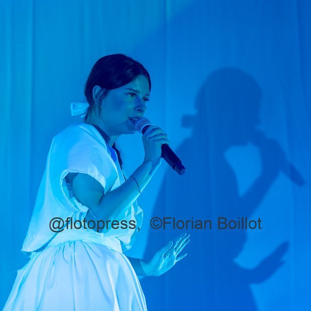A singer performs on stage, illuminated by blue lighting, holding a microphone.