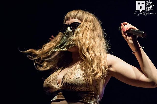 A woman with a gold mask performs on stage with a microphone in hand during a live concert.