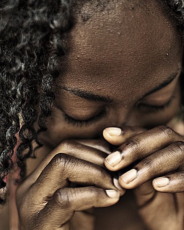A woman with dark skin is shown in a close-up with her eyes closed and hands clasped near her face.