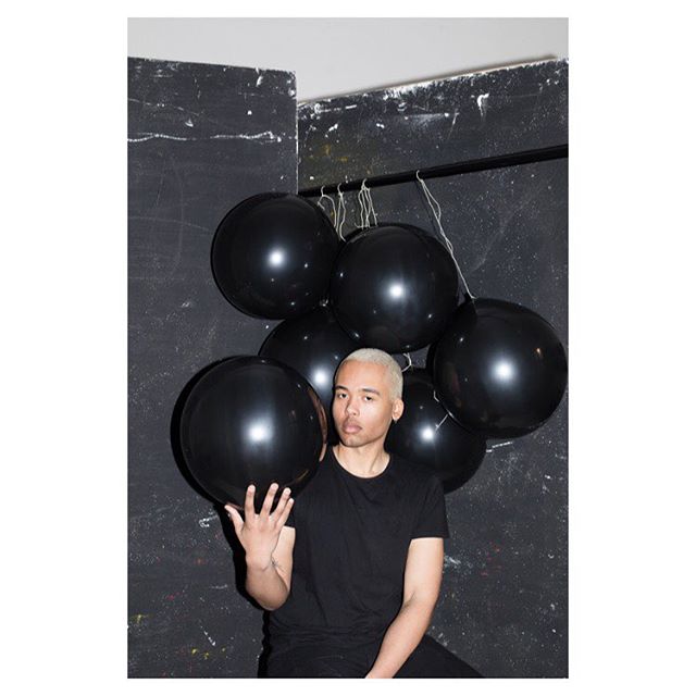 A man poses with a bunch of black balloons in a studio, against a textured background.