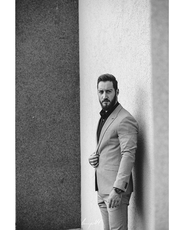 A bearded man in a suit stands confidently against a wall in a black and white portrait.