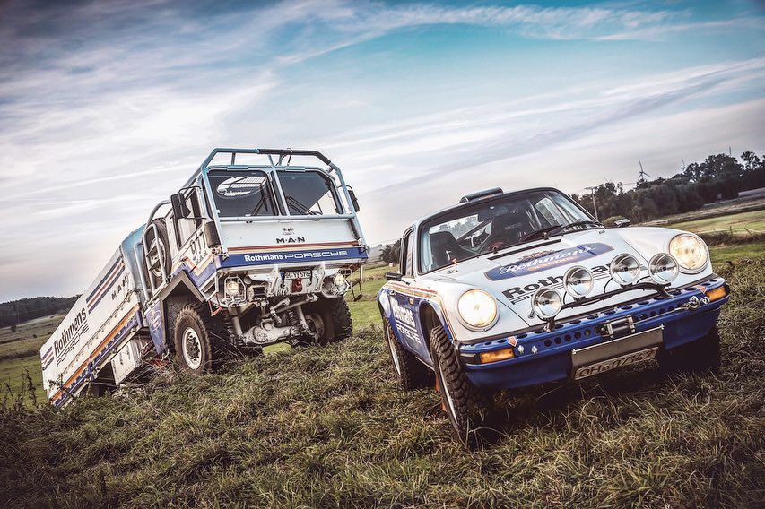 An off-road Porsche and a MAN truck with Rothmans livery are parked on a grassy field under a cloudy sky.