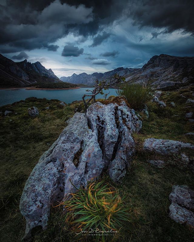 A moody mountain lake scene with dramatic clouds and a rock formation in the foreground.