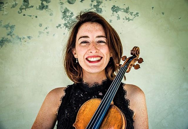 A smiling woman holds a violin indoors, radiating joy and passion for music.